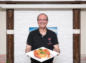 Der lächelnde Besitzer des griechischen Lokals hält einen Teller Akastomakaronada, ein griechisches Nudelgericht ins Bild.